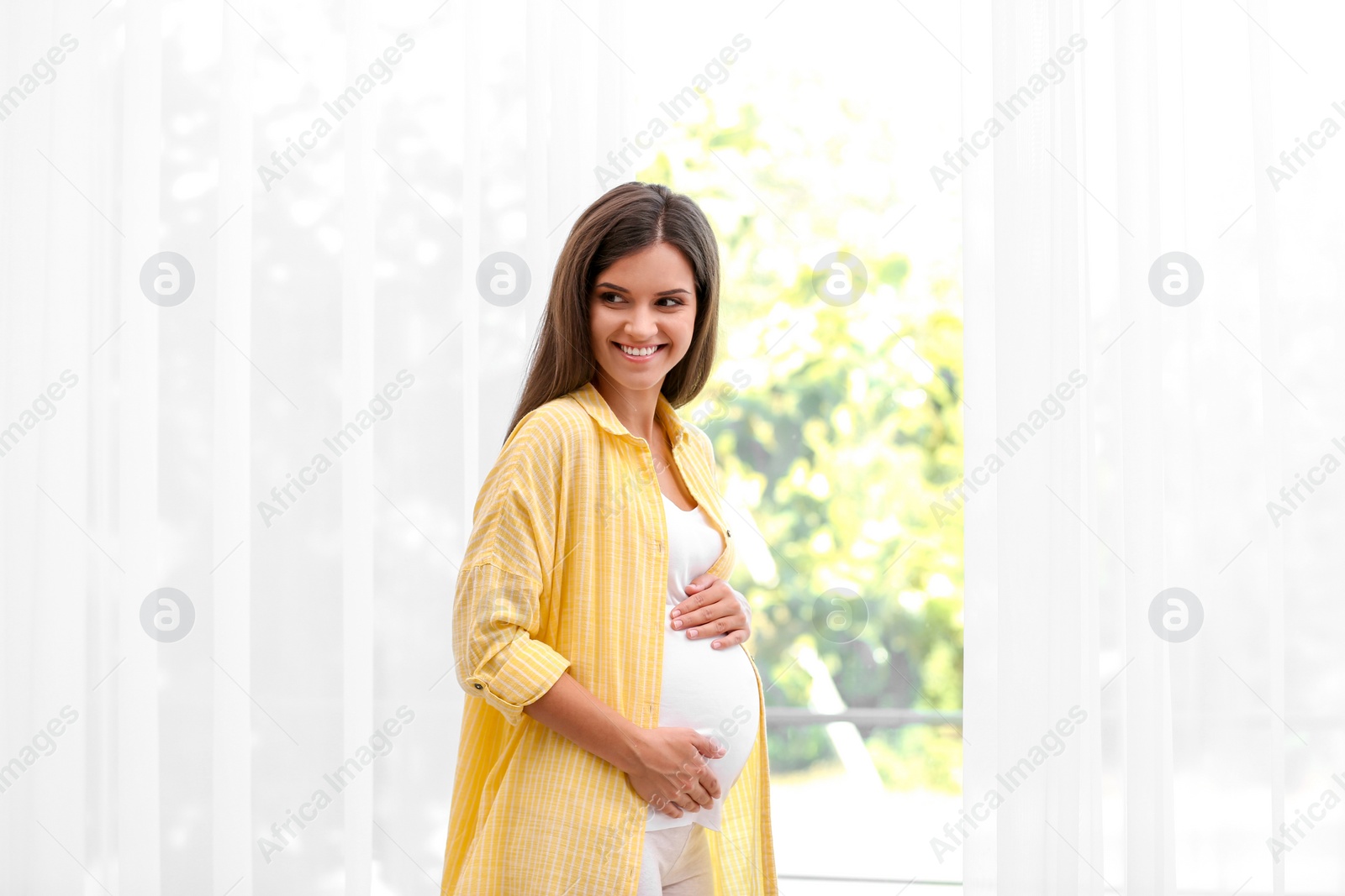 Photo of Happy pregnant woman standing near window at home