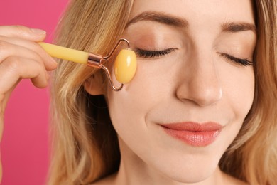 Young woman using natural jade face roller on pink background, closeup