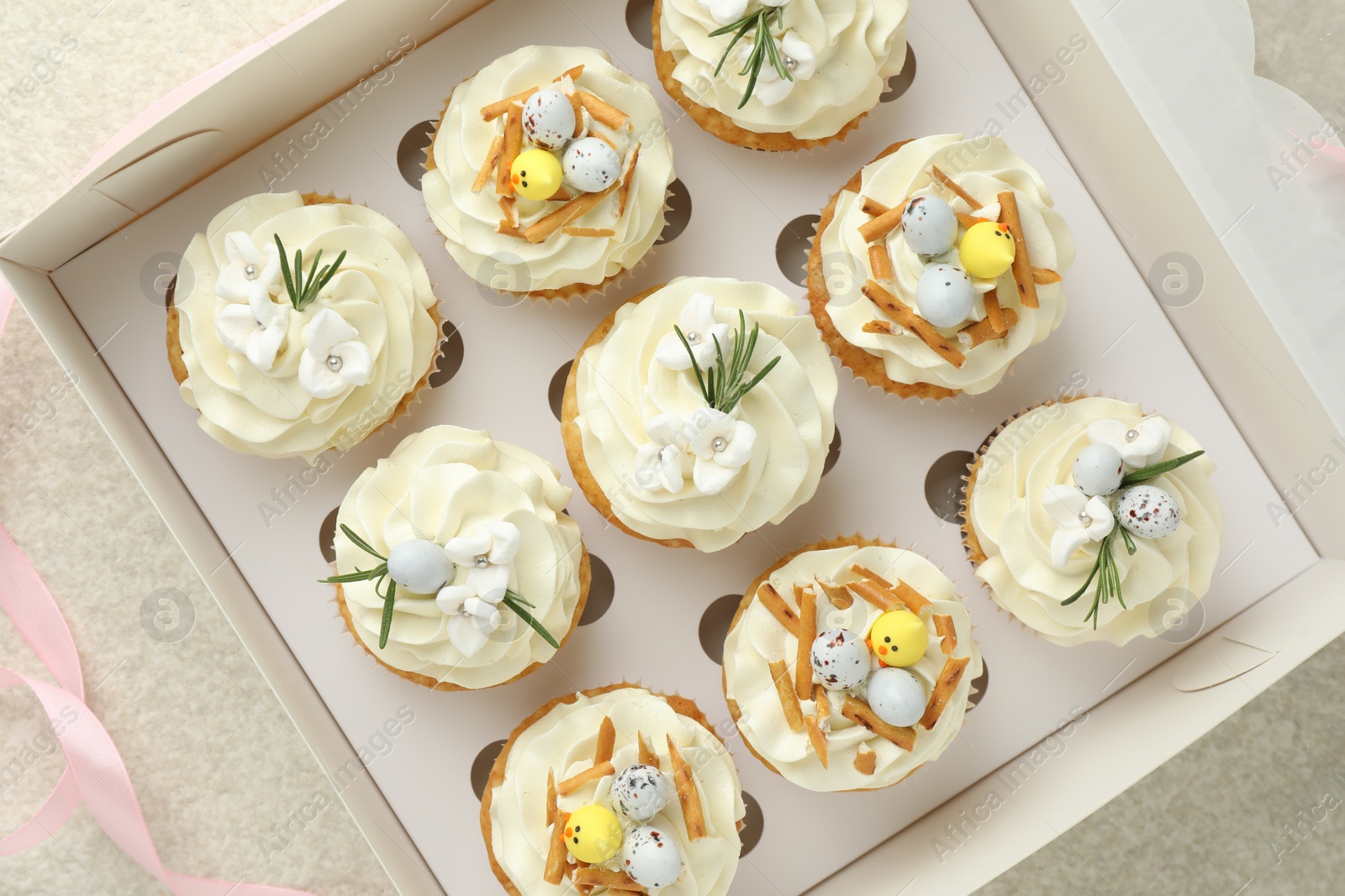 Photo of Tasty Easter cupcakes in box and pink ribbon on beige table, flat lay