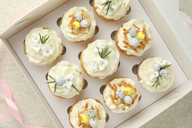 Tasty Easter cupcakes in box and pink ribbon on beige table, flat lay