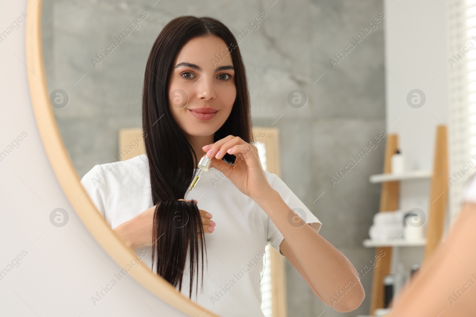 Photo of Beautiful woman applying hair serum in bathroom, reflection in mirror. Cosmetic product