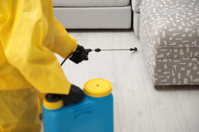 Photo of Pest control worker spraying pesticide under furniture indoors, closeup