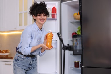 Photo of Smiling food blogger with bottle of juice recording video in kitchen