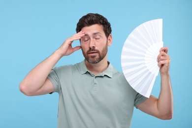 Unhappy man with hand fan suffering from heat on light blue background