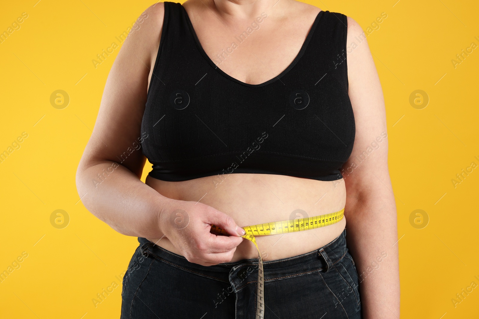 Photo of Overweight woman measuring waist with tape on orange background, closeup