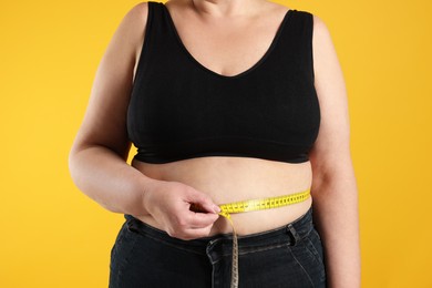 Overweight woman measuring waist with tape on orange background, closeup