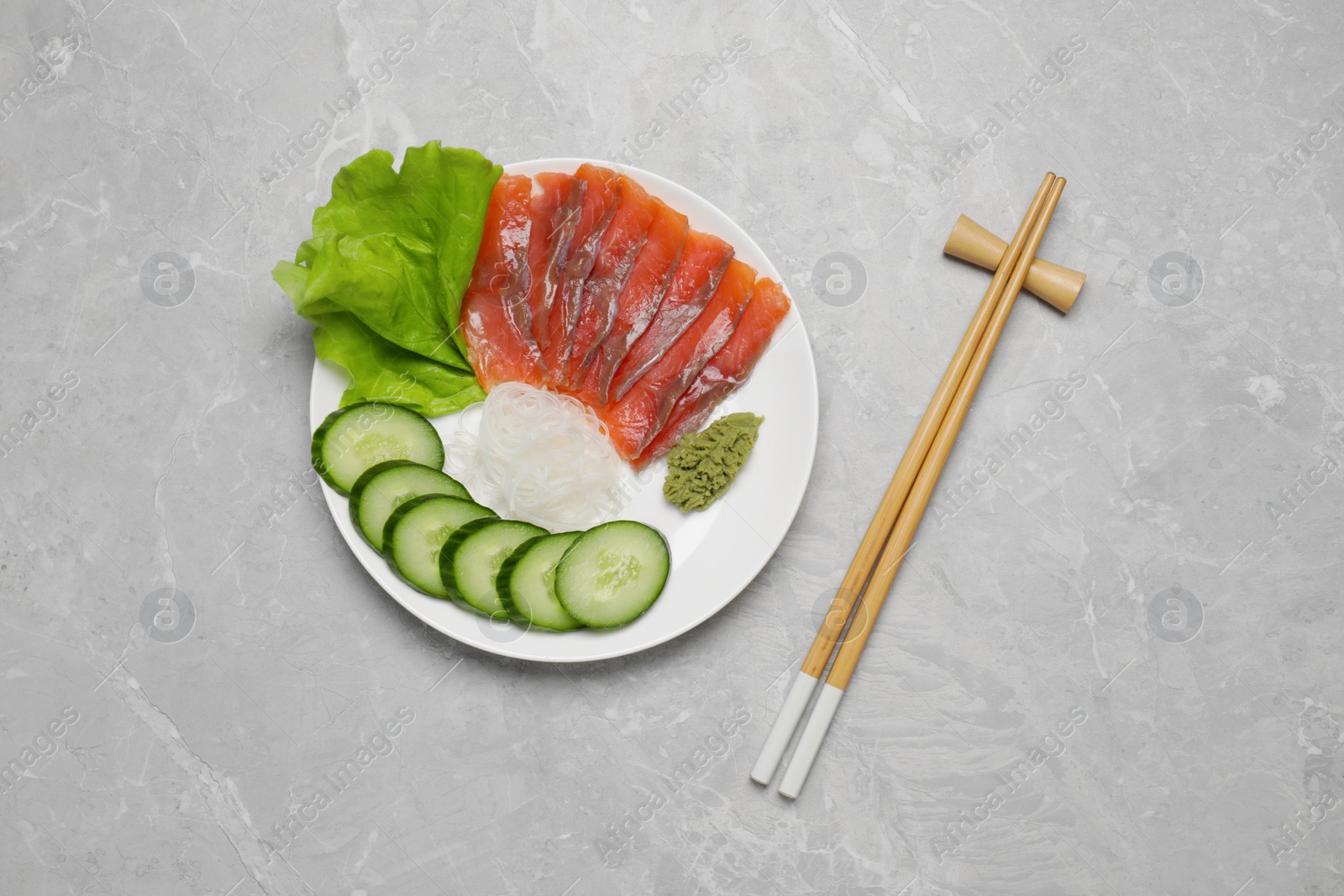 Photo of Sashimi set (salmon slices) with cucumber, greens, vasabi, funchosa and chopsticks on light grey table, flat lay