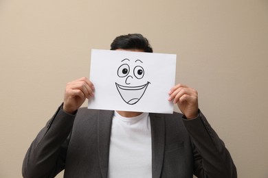 Photo of Man hiding emotions using card with drawn smiling face on beige background