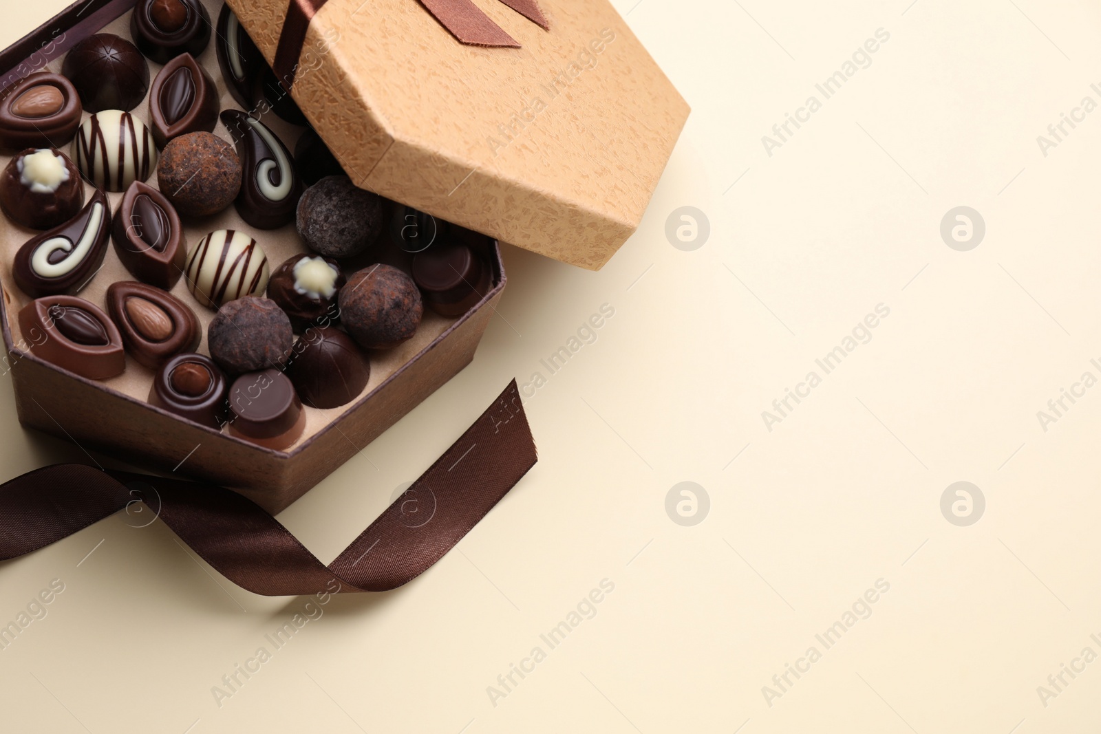 Photo of Open box of delicious chocolate candies and brown ribbon on beige background, flat lay. Space for text
