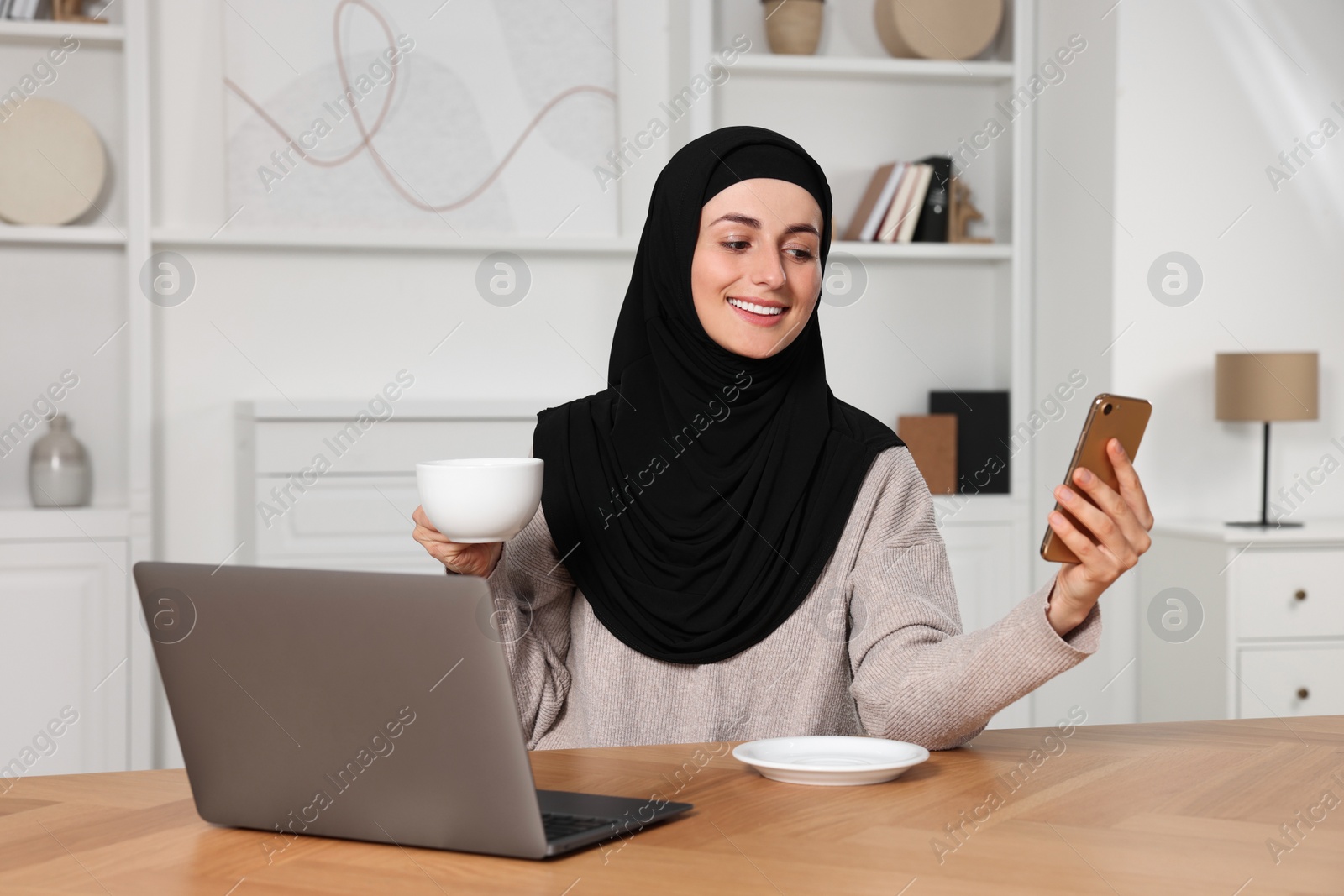 Photo of Muslim woman using smartphone near laptop at wooden table in room