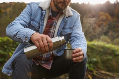 Photo of Man pouring hot drink from metallic thermos into cup lid in nature, closeup