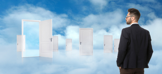 Image of Young man standing in front of many similar doors against blue sky. Choice concept