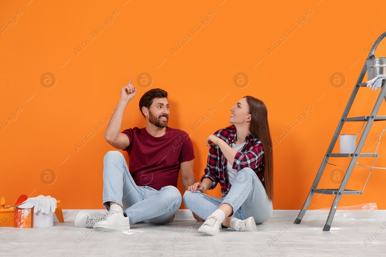 Photo of Man pointing upwards and woman sitting on floor near freshly painted orange wall indoors. Interior design