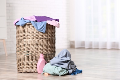 Photo of Wicker laundry basket with dirty clothes and detergent on floor indoors. Space for text