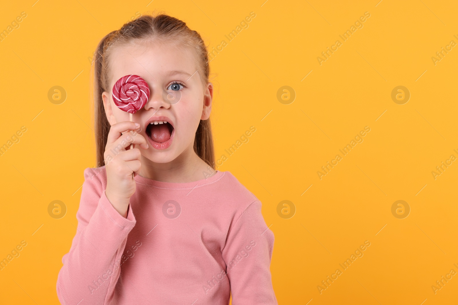 Photo of Emotional little girl covering eye with bright lollipop swirl on orange background, space for text