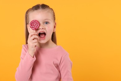 Photo of Emotional little girl covering eye with bright lollipop swirl on orange background, space for text