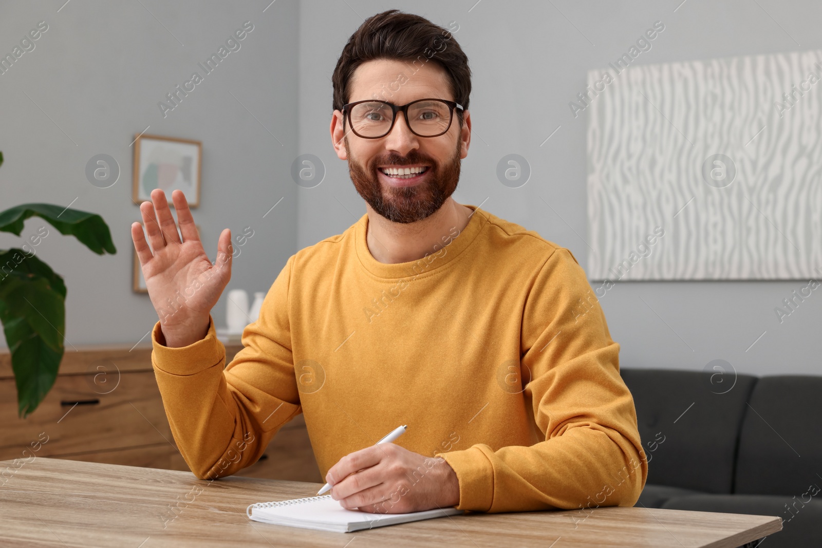 Photo of Man in glasses greeting someone at wooden table indoors