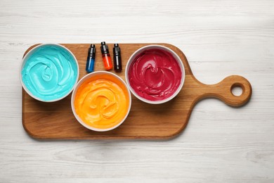 Bowls of different cream with food coloring and bottles of bright liquid on white wooden table, top view