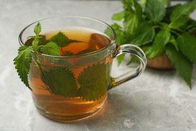 Glass cup of aromatic nettle tea and green leaves on light grey table, closeup