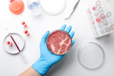 Photo of Scientist holding Petri dish with cultured meat above white lab table, top view