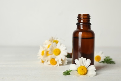 Photo of Chamomile flowers and cosmetic bottle of essential oil on wooden table against light background. Space for text