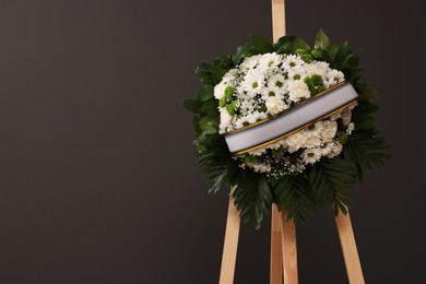 Photo of Funeral wreath of flowers with ribbon on wooden stand against grey background, space for text