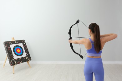 Photo of Woman with bow and arrow aiming at archery target indoors, back view