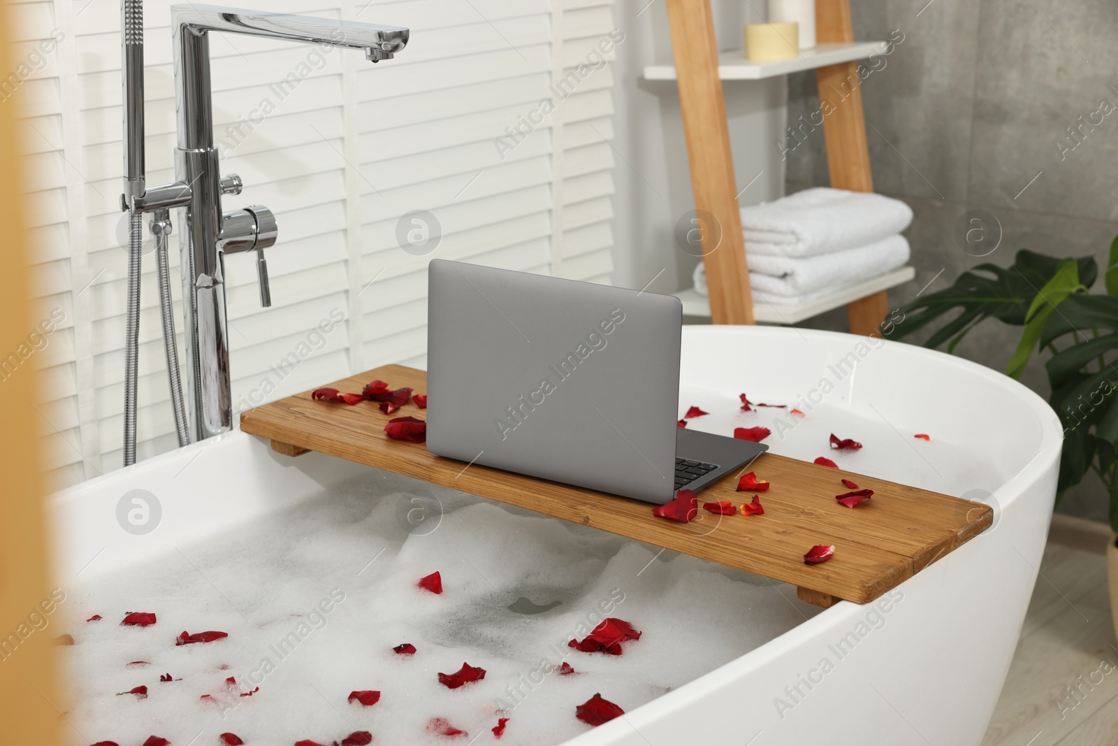 Photo of Wooden board with laptop and rose petals on bath tub in bathroom