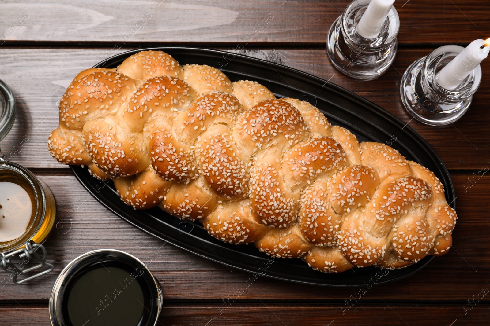Photo of Homemade braided bread with sesame seeds, goblet, honey and candles on wooden table, flat lay. Traditional Shabbat challah