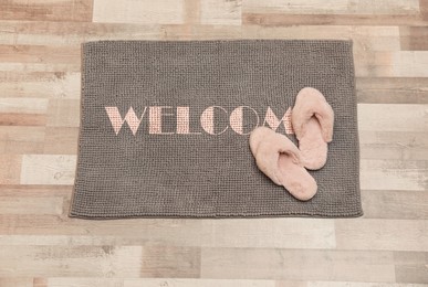 Image of Door mat with word WELCOME and fluffy slippers on floor, top view