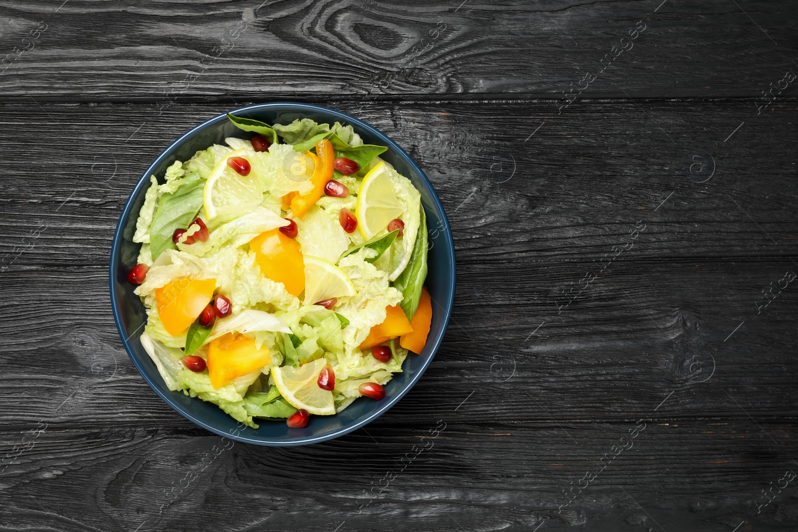 Photo of Delicious salad with Chinese cabbage, lemon, persimmon and pomegranate seeds on black wooden table, top view. Space for text