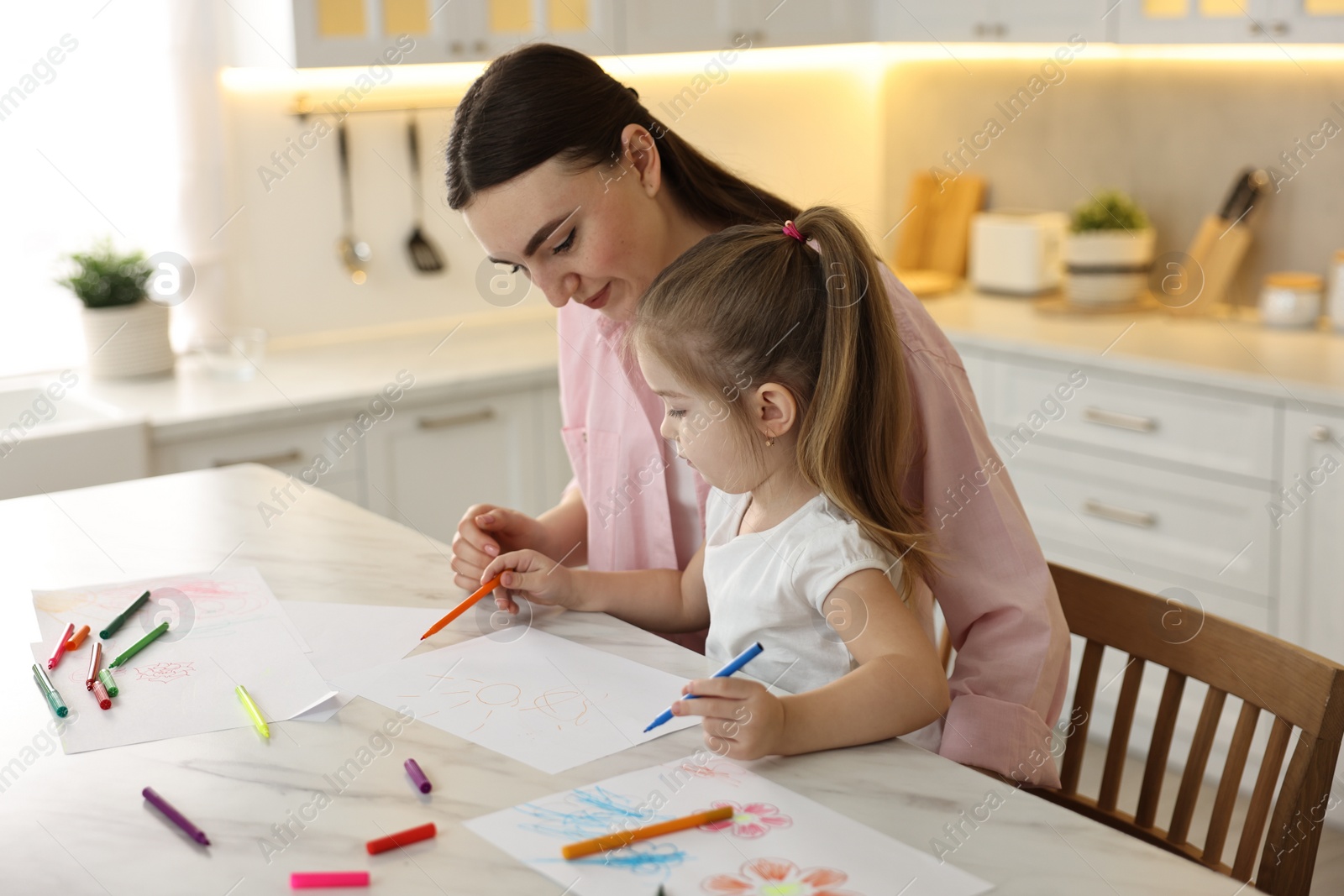 Photo of Mother and her little daughter drawing with colorful markers at table in kitchen