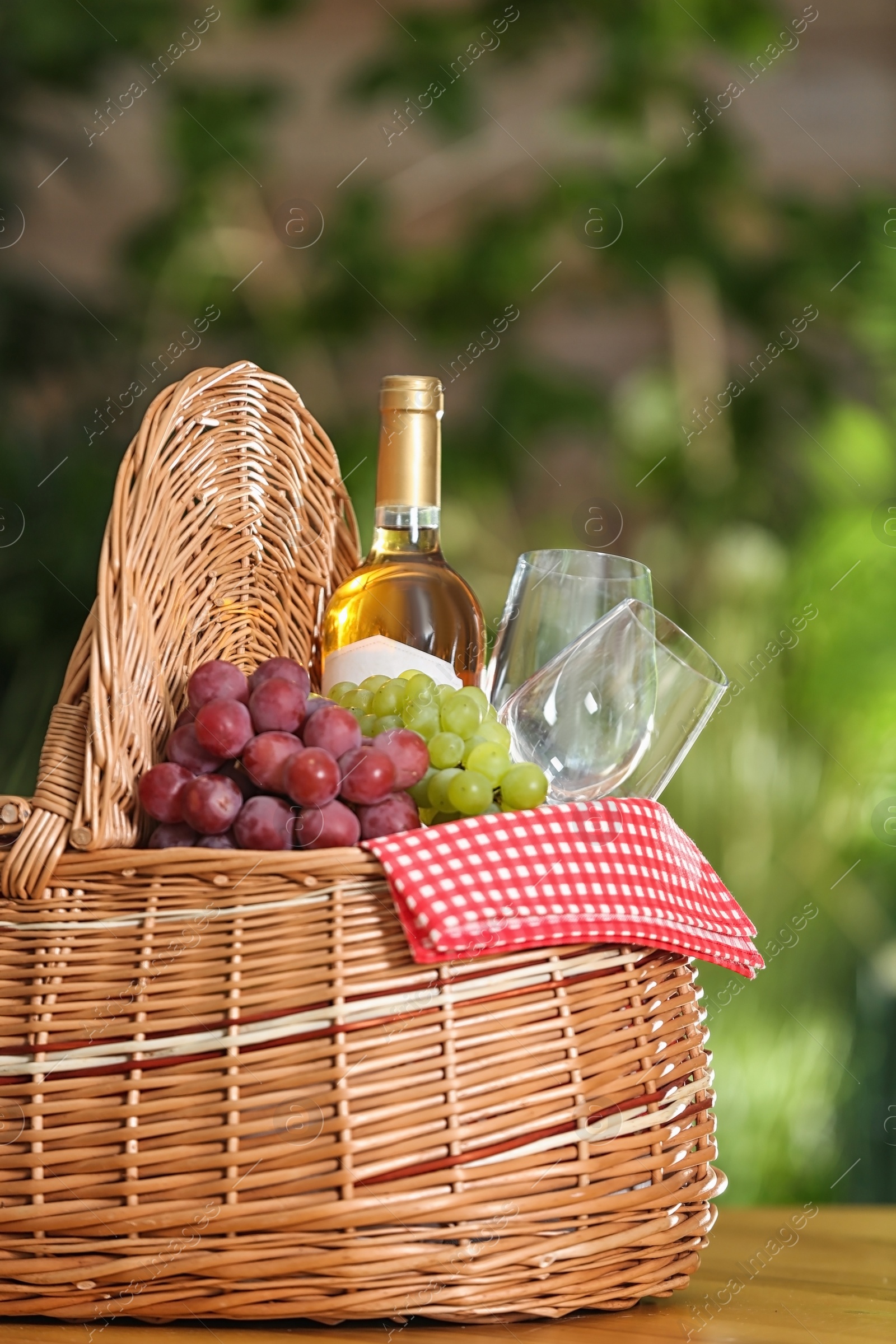 Photo of Picnic basket with wine, glasses and grapes on wooden table against blurred background, space for text