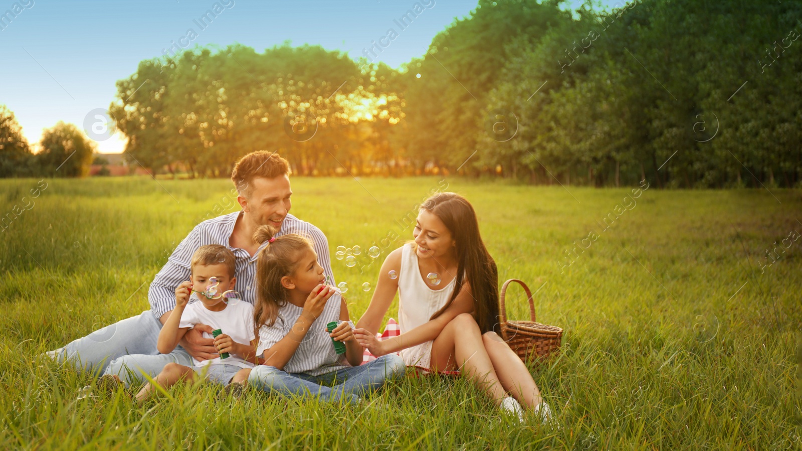 Image of Happy family having picnic and blowing soap bubbles in park at sunset