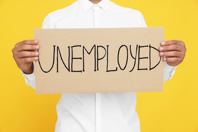 Young man holding sign with word Unemployed on yellow background, closeup