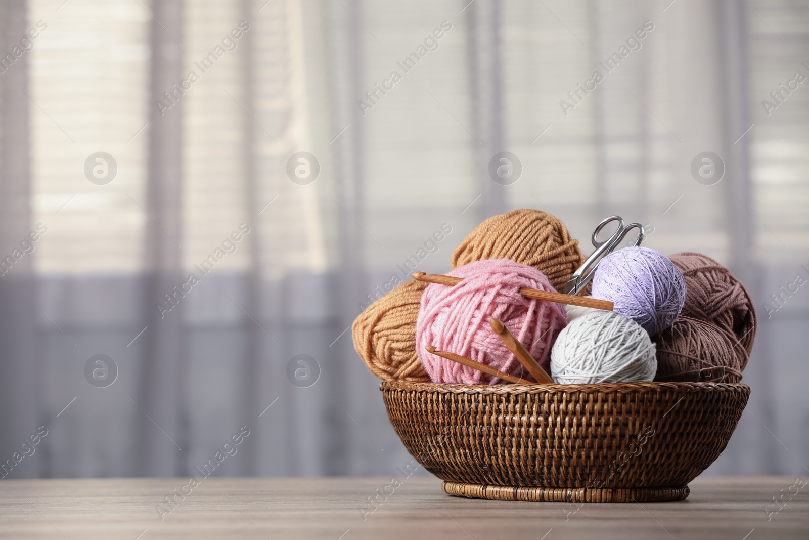 Photo of Wicker basket with clews of colorful knitting threads, crochet hooks and scissors on wooden table indoors. Space for text