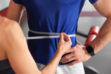 Photo of Trainer measuring man`s waist with tape in gym, closeup