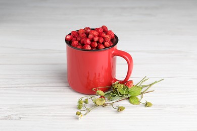 Fresh wild strawberries in mug and green stems on white wooden table