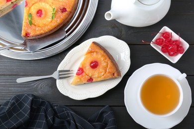 Photo of Delicious cut pineapple pie with cherry and tea served on black wooden table, flat lay