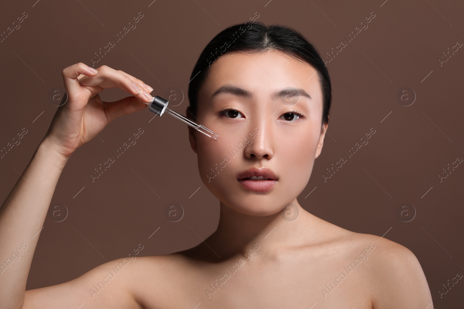 Photo of Beautiful young woman applying cosmetic serum onto her face on brown background