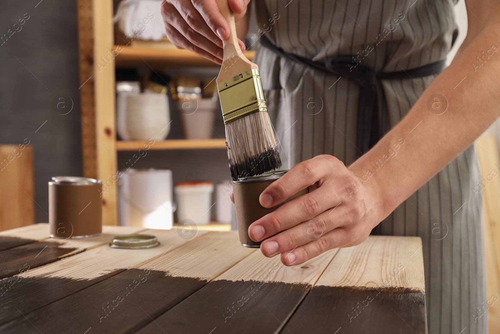 Photo of Man dipping brush into can of wood stain at wooden surface indoors, closeup