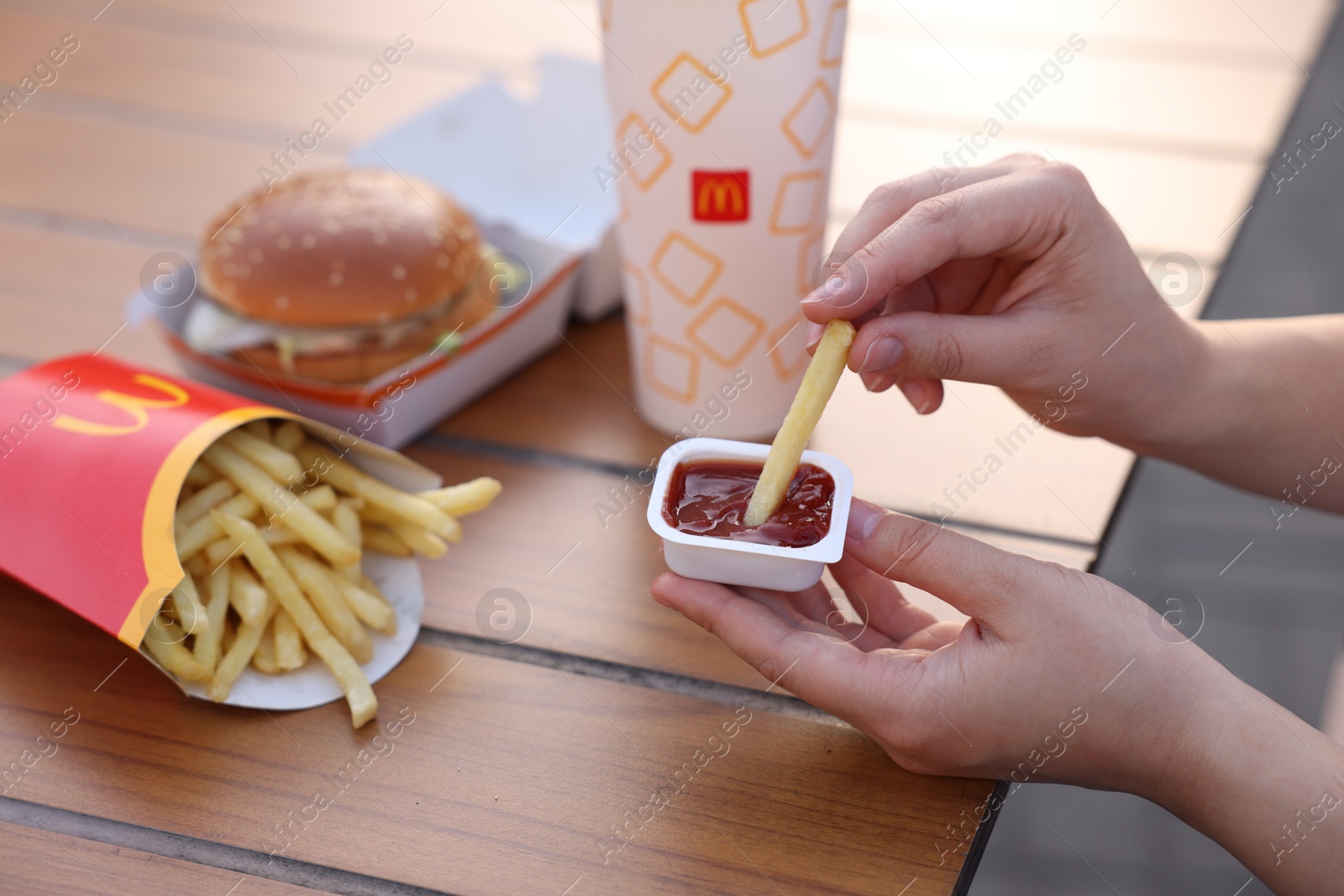 Photo of Lviv, Ukraine - September 26, 2023: Woman dipping McDonald's french fry into sauce at wooden table outdoors, closeup