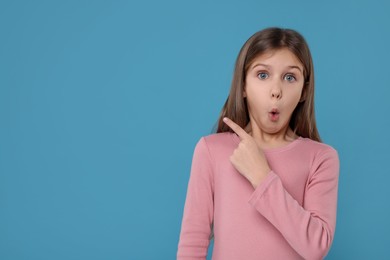 Photo of Surprised girl pointing at something on light blue background. Space for text