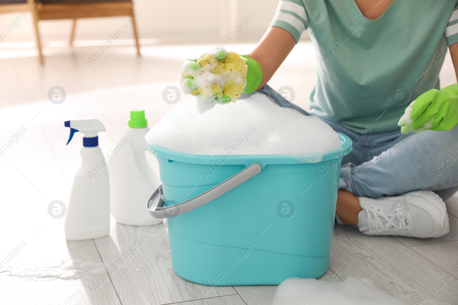 Photo of Woman holding sponge with foam over bucket indoors, closeup. Cleaning supplies