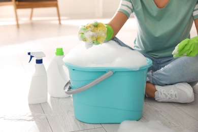 Woman holding sponge with foam over bucket indoors, closeup. Cleaning supplies