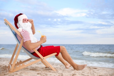 Photo of Santa Claus with cocktail relaxing in chair on beach, space for text. Christmas vacation