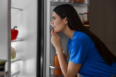 Thoughtful young woman looking into modern refrigerator at night