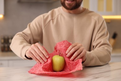 Man packing fresh apple into beeswax food wrap at light marble table in kitchen, closeup