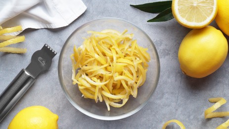 Photo of Zester, fresh lemons and bowl with pieces of peel on grey table, flat lay