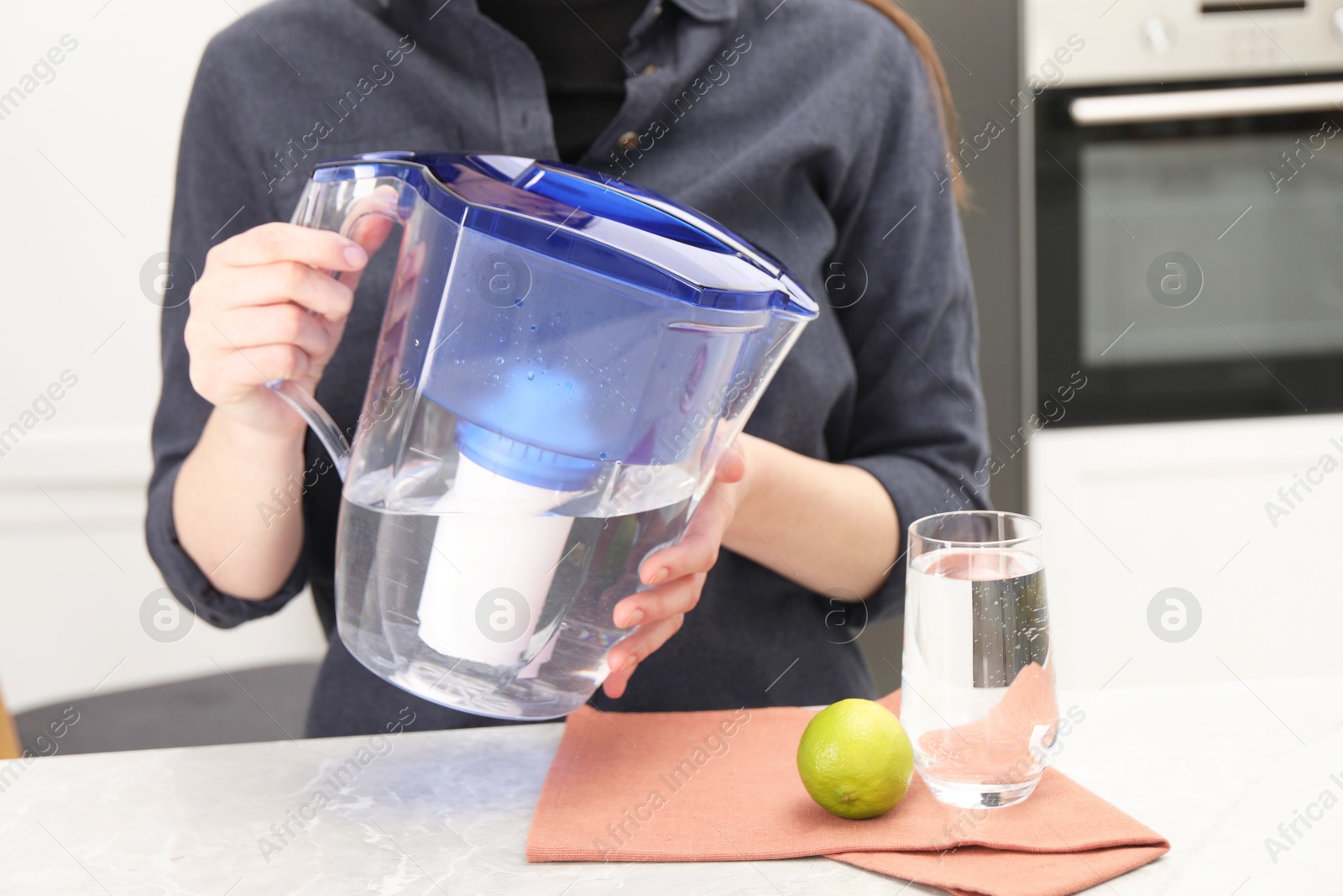 Photo of Woman with water filter jug at light marble table in kitchen, closeup